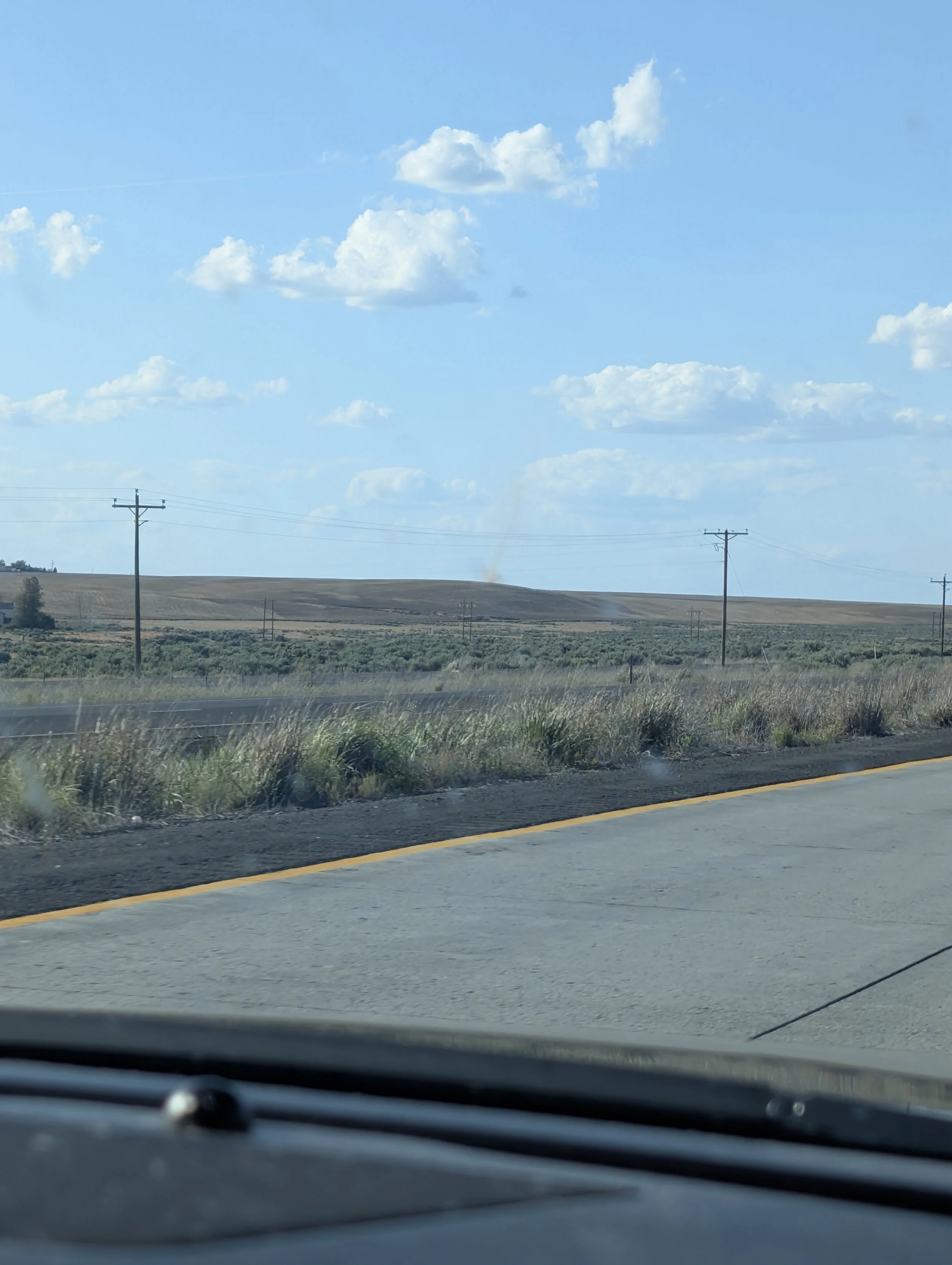 View from inside a moving vehicle showing a rural highway with power lines, a distant plume of smoke rising from a hill, and a cloudy blue sky.