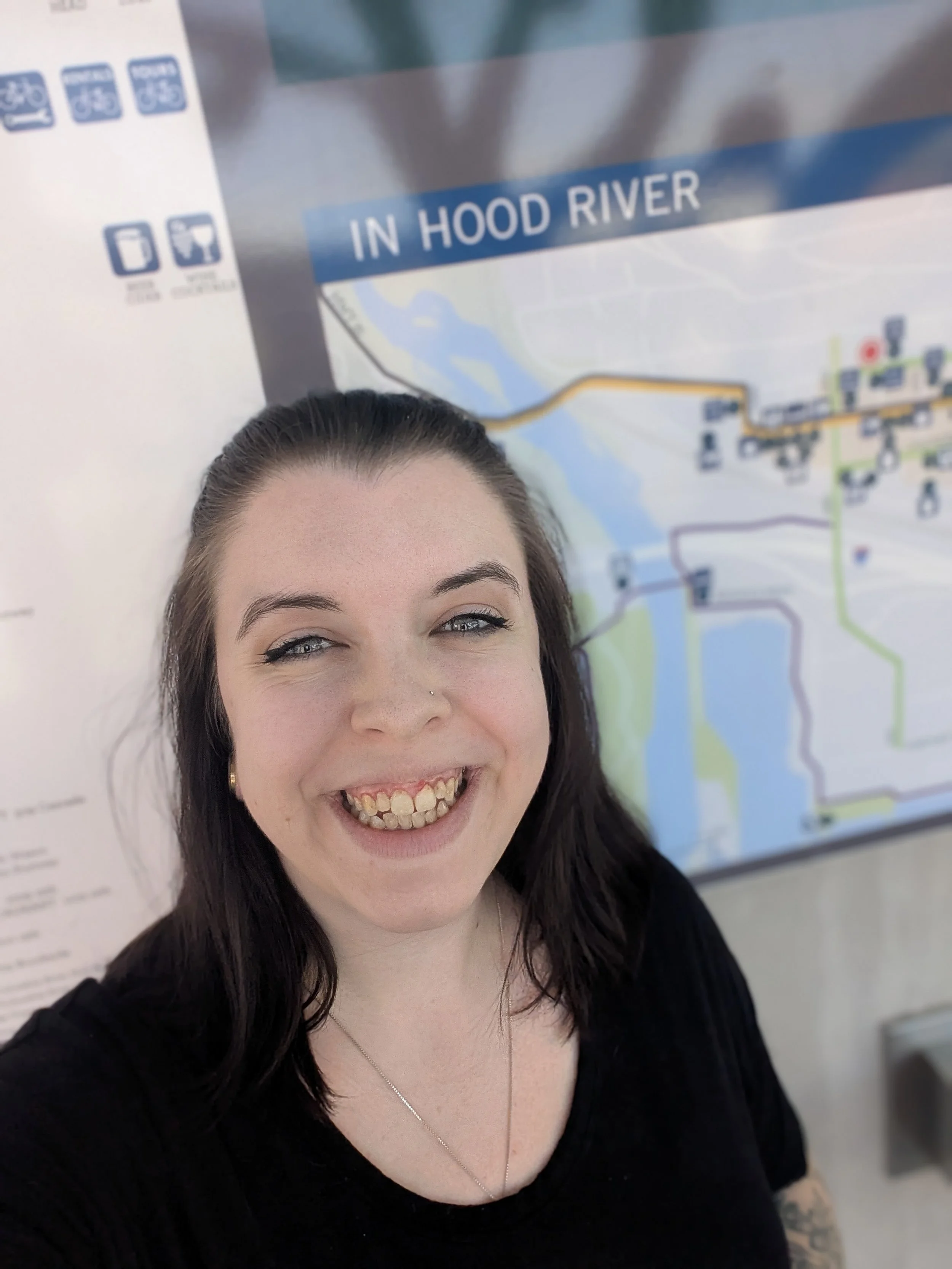 A woman with dark brown hair, eyes half-closed, and a broad smile showing her teeth, taking a selfie against a colorful map background in an indoor setting.