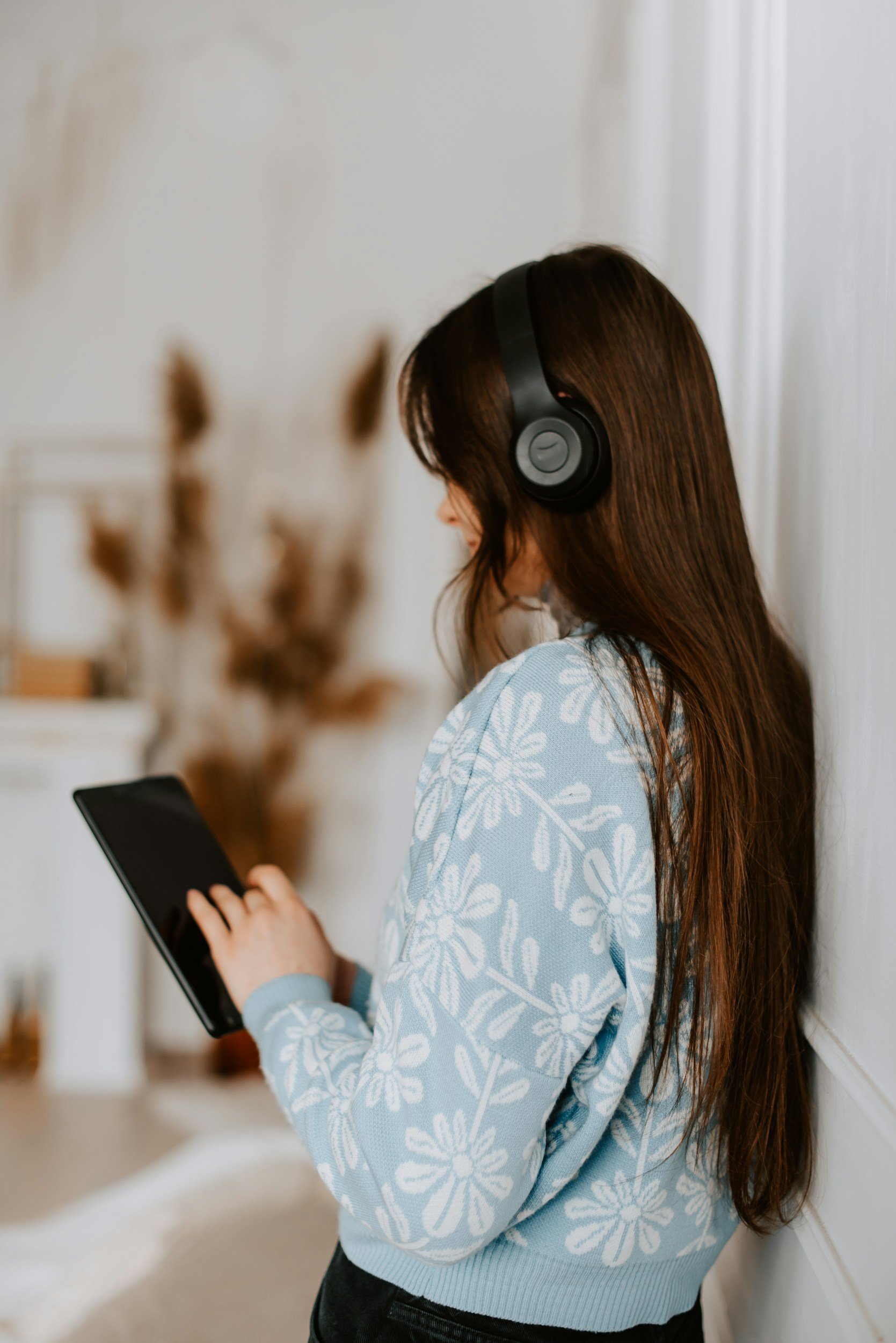 A young woman with long dark hair wearing headphones and a light blue sweater with white floral patterns, standing indoors and looking at a tablet device.