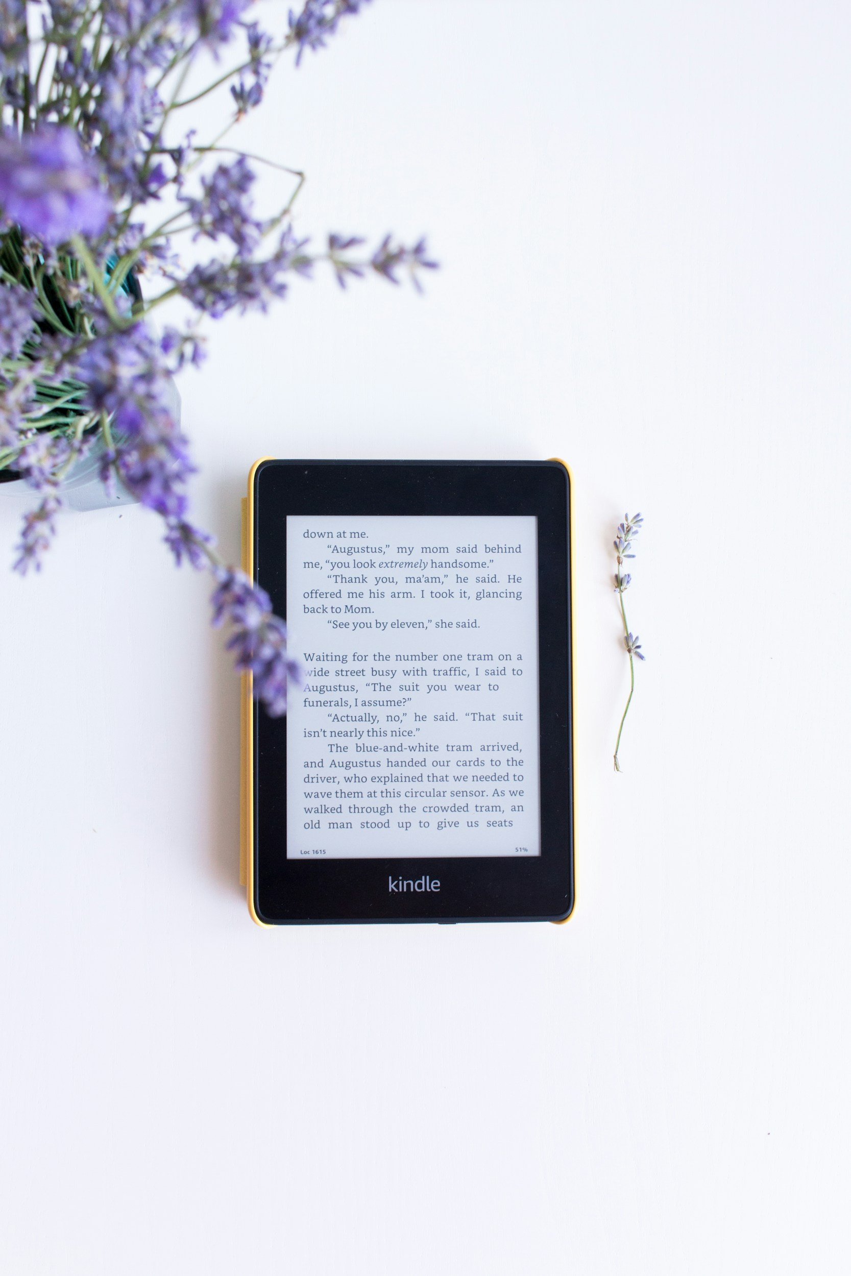 A Kindle e-reader displaying text, positioned on a white surface. A sprig of lavender flowers to the right and a vase of purple lavender flowers on the left.