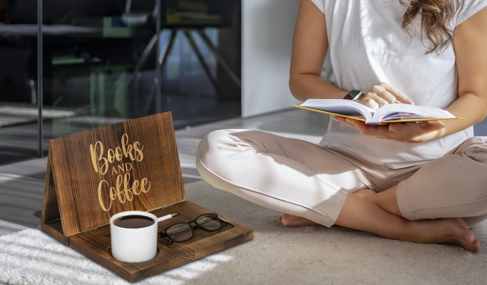 Person sitting cross-legged on the floor reading a book, with a wooden sign saying "Books and Coffee," a mug of coffee, and a pair of glasses on the floor next to her.