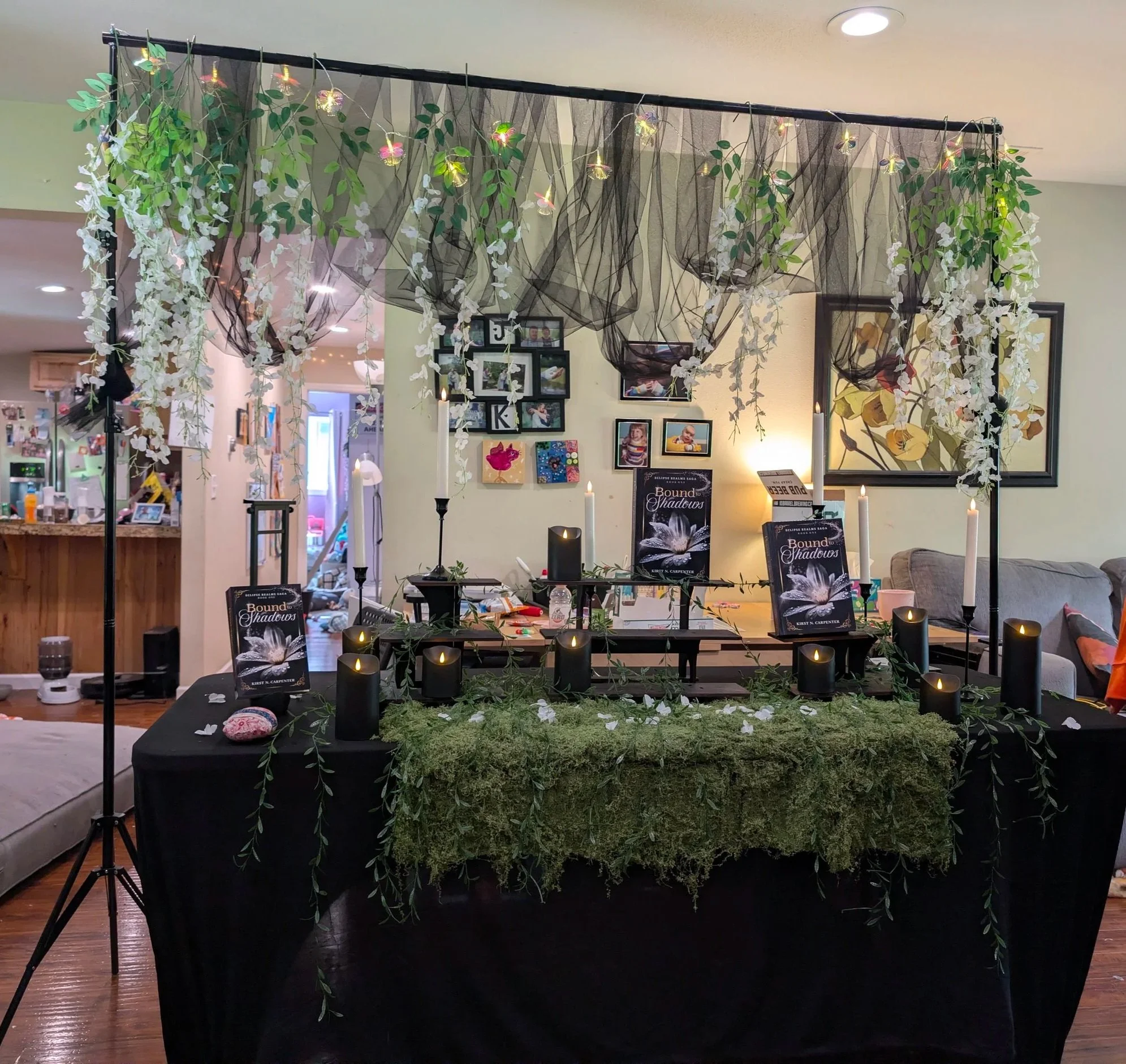 Decorative indoor setup with black candles, greenery, and white flowers on a table, wall art, and framed photographs in the background.