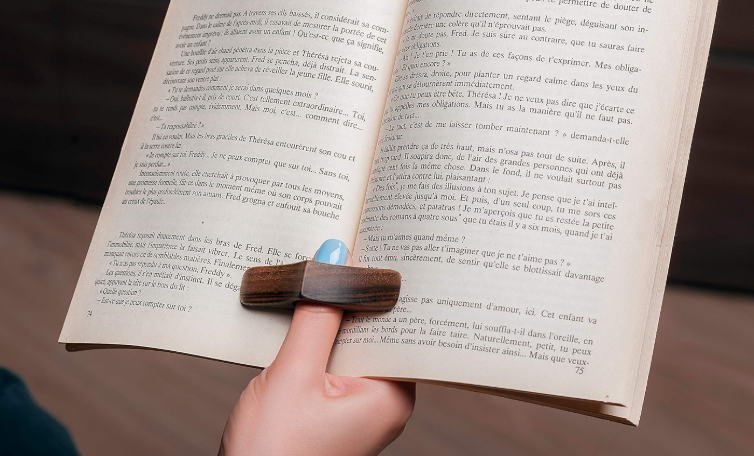 A person is holding an open book with a wooden bookmark, positioned over the middle of the pages, on a wooden surface.