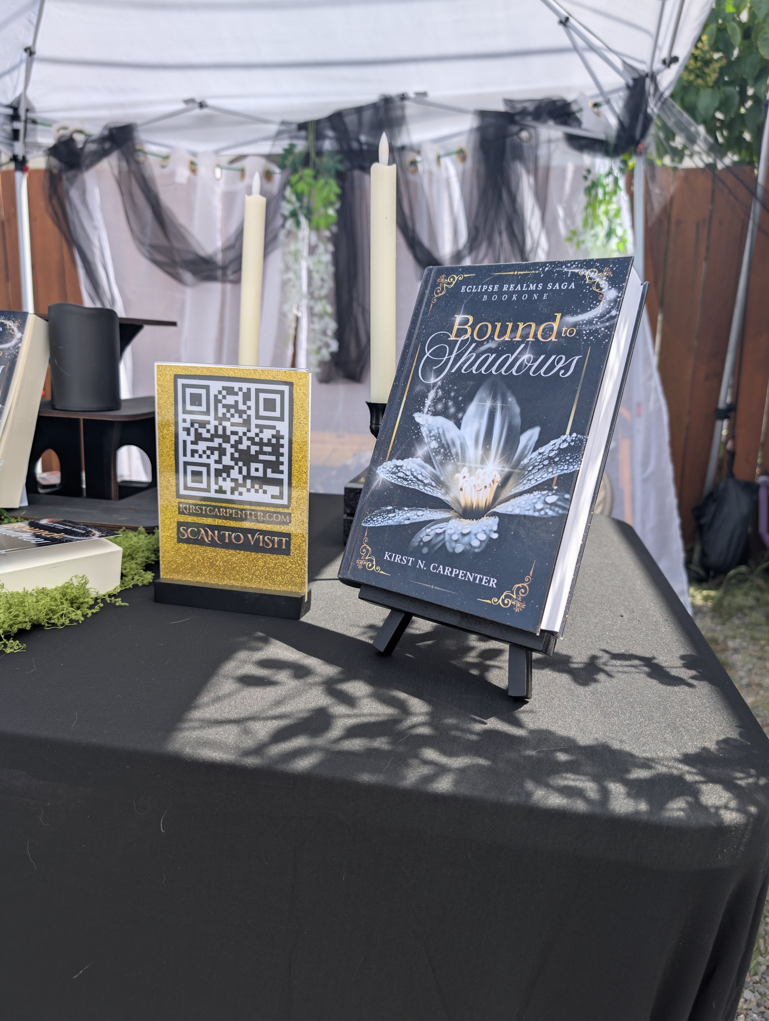 Table display about the book titled 'Bound to Shadows' by Kirst N. Carpenter from the Eclipse Realms Saga, featuring a sign with a QR code for website visit, tall white candles, and decorative elements, set under a white canopy with hanging black and white fabric and greenery.