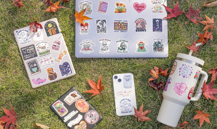 Laptop, phone, mug, and four sheets of stickers with book and library-themed designs, scattered on grass with fall leaves.