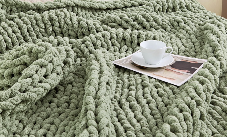 A white coffee cup and saucer with a magazine resting on a chunky, textured, light green blanket.