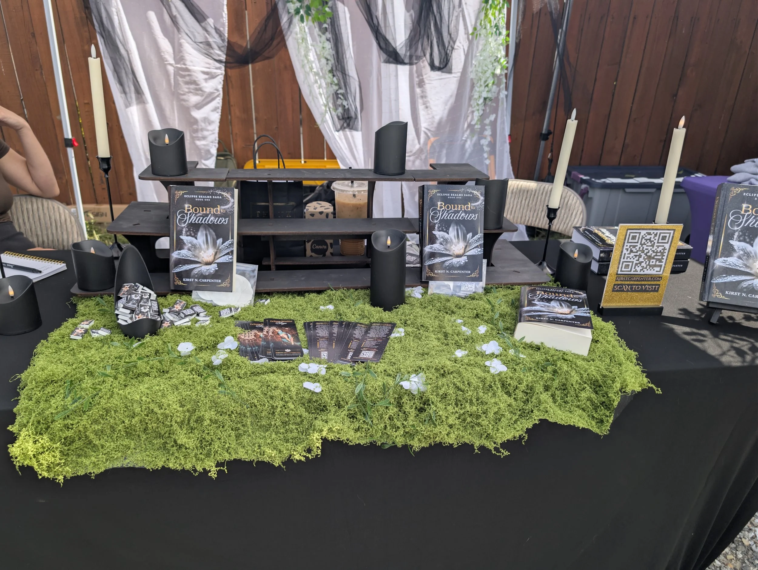 A display table with books titled 'Bound to Shadows' by Kirst N. Carpenter, featuring black candles, pamphlets, a QR code, and decorative elements, set on a green moss covering with a wooden fence and white curtains in the background.