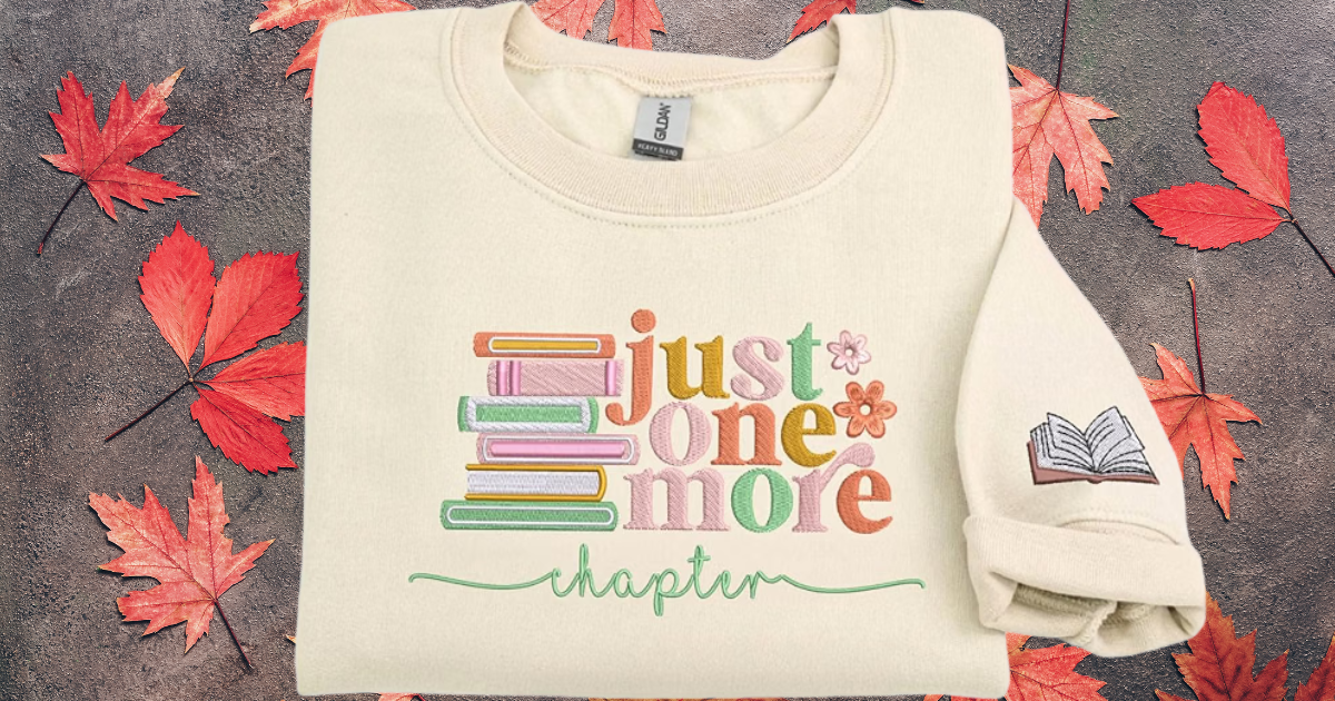 Cream-colored sweatshirt with the phrase "just one more chapter" and a stack of books graphic printed on it, surrounded by red fall leaves
