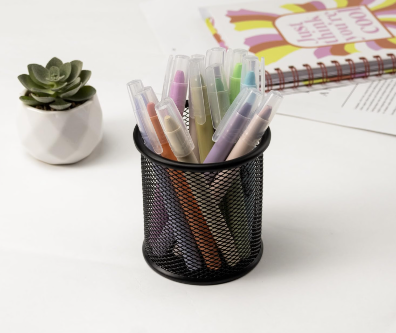 A black mesh pen holder filled with pastel-colored highlighters and markers on a white desk.