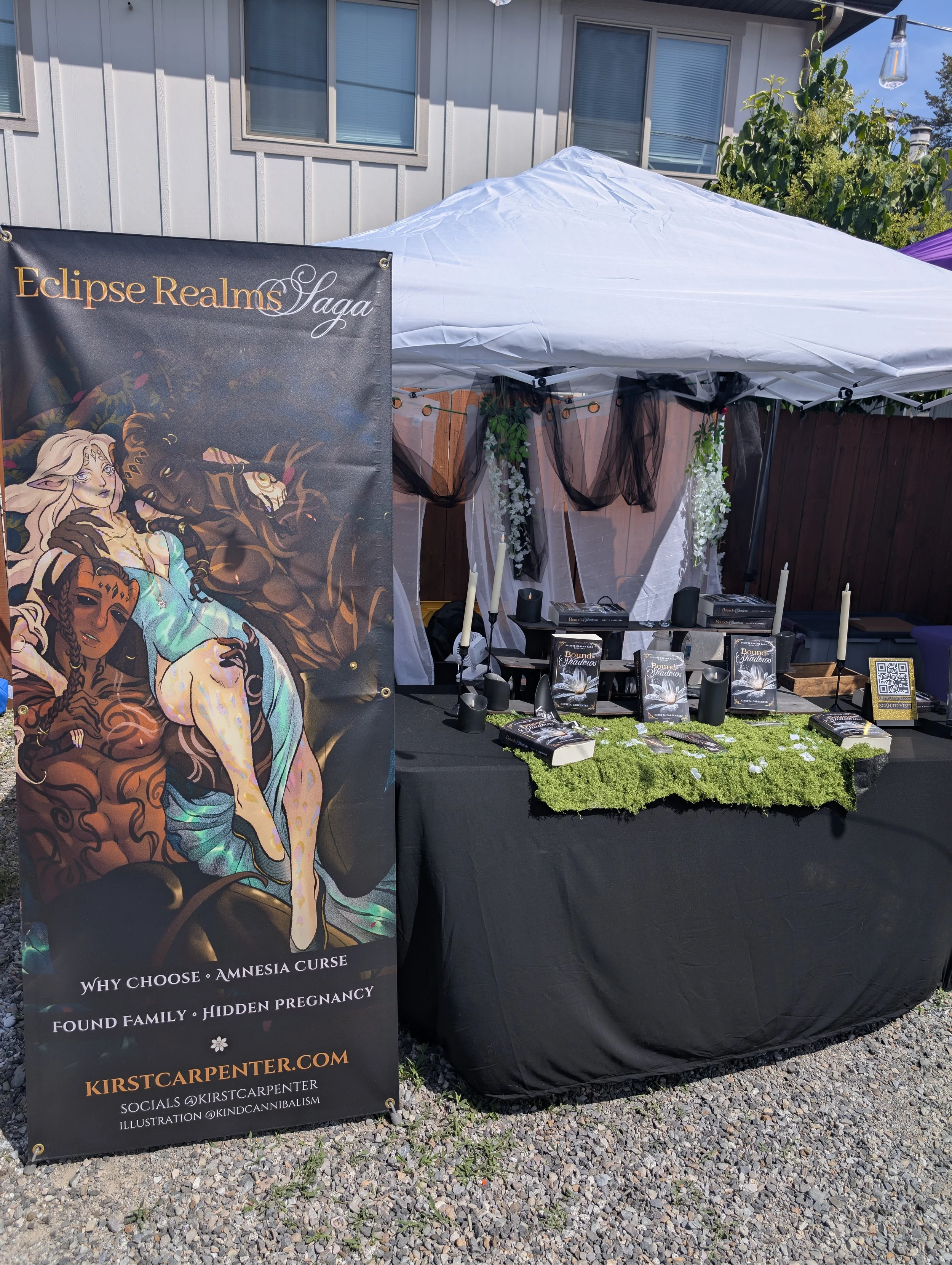 Outdoor booth with a black tablecloth, books, and candles, next to a banner featuring fantasy artwork, with a white canopy tent, decorative black and white curtains, and some greenery in the background.