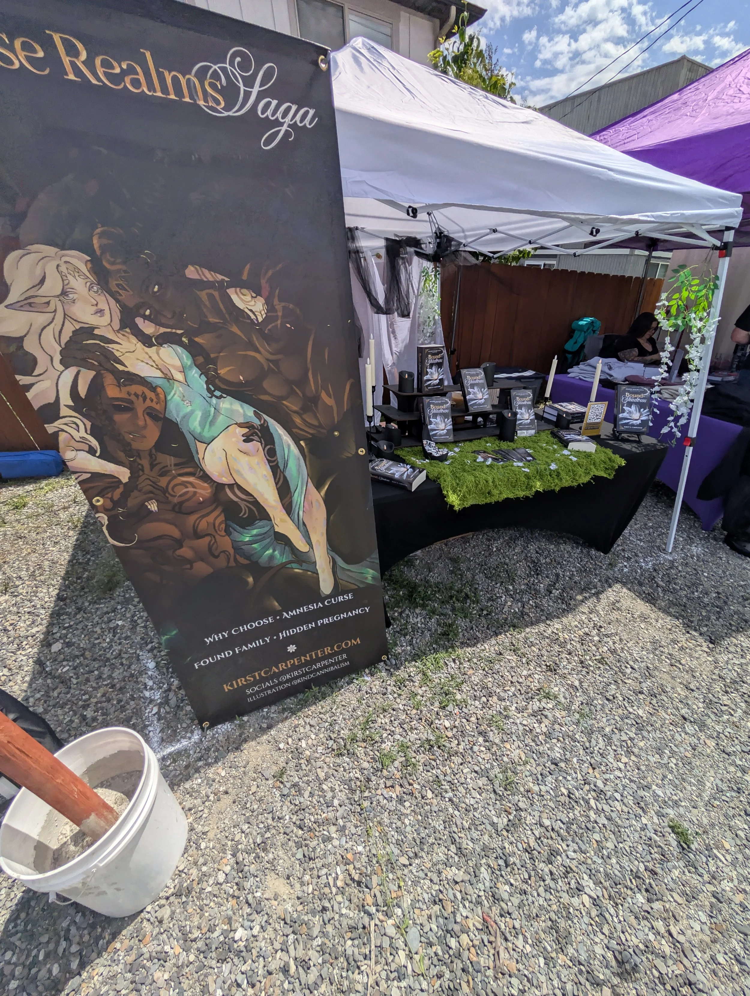 A booth at an outdoor market shows a black banner with artwork of a woman with long, flowing hair and fantasy-style makeup. The banner advertises services related to choosing, amnesia curses, and hidden pregnancy, with contact information and a website. The booth has a table with books, pamphlets, and decorative items, including candles, and is covered by white and purple tents. There are people and other market stalls visible in the background, and the ground is gravel.
