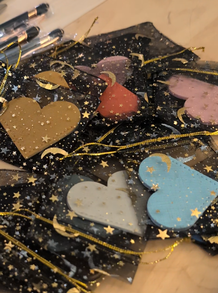 Assorted decorative heart and moon-shaped cutouts in pastel pink, blue, white, beige, and red, decorated with gold stars and moons, inside a transparent bag with gold star embellishments.