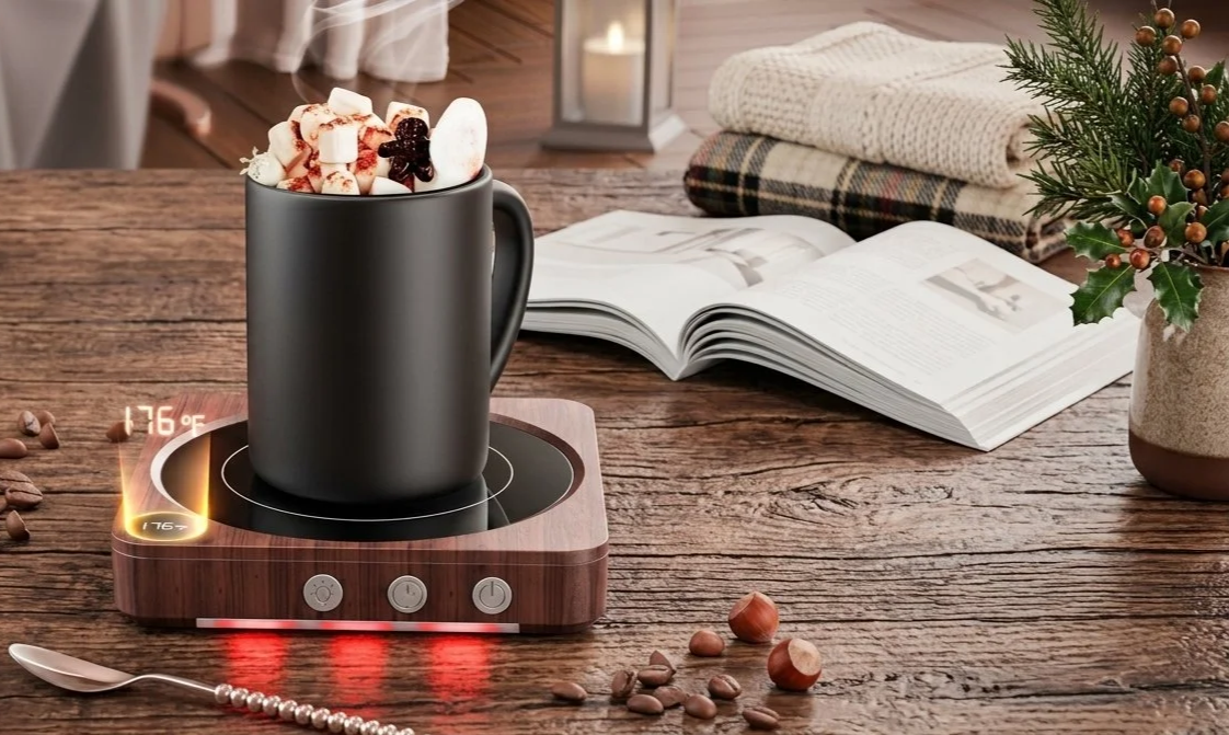 A mug filled with hot chocolate and topped with marshmallows on a wooden table with a digital hot chocolate warmer, an open magazine, folded blankets, hazelnuts, coffee beans, a necklace, and a small potted holiday plant.