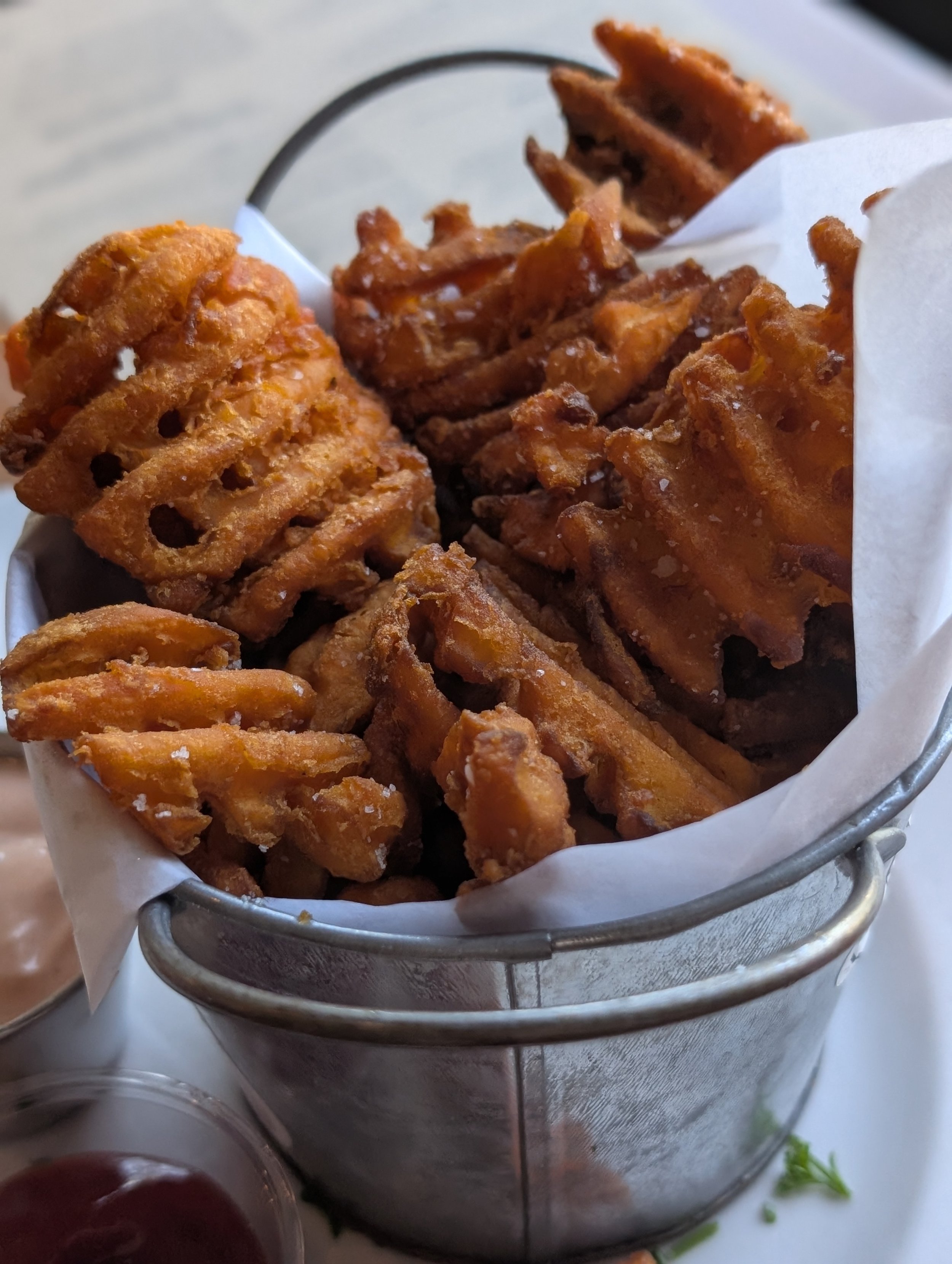 Fried lotus root chips in a small metal basket lined with white paper, placed on a metal tray, with a small bowl of red sauce nearby.