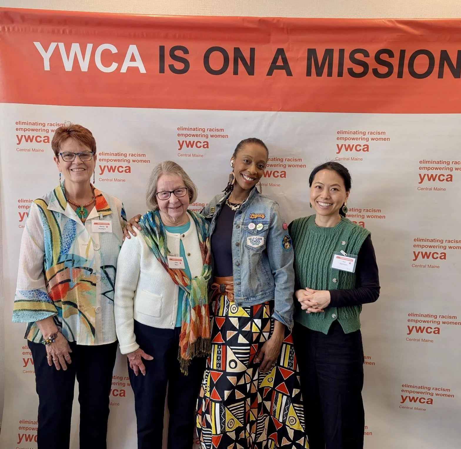 Four women standing in front of a YWCA banner that reads 'YWC A is on a mission,' smiling and posing for the photo.
