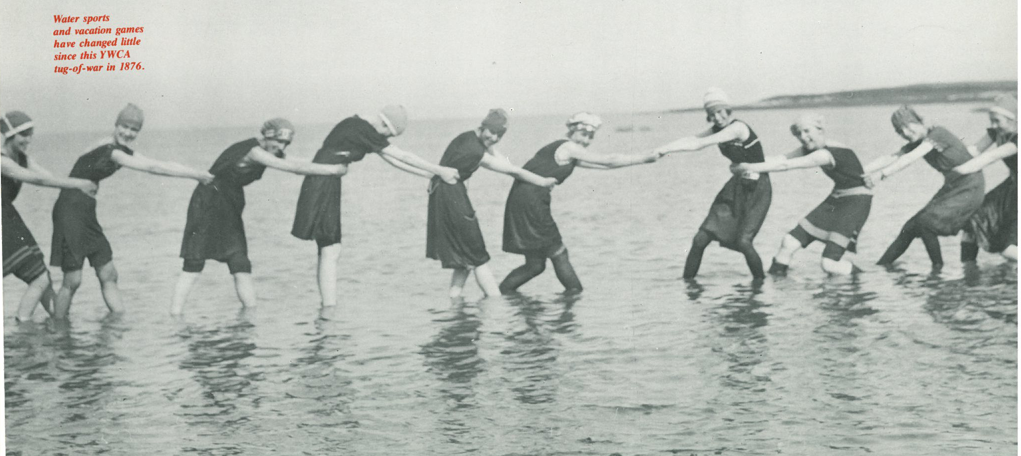 Black and white photo of women in vintage swimsuits and bathing caps playing tug-of-war in shallow water on the beach.