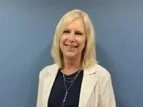A smiling woman with blonde hair wearing a light-colored blazer over a dark top, standing against a blue background.