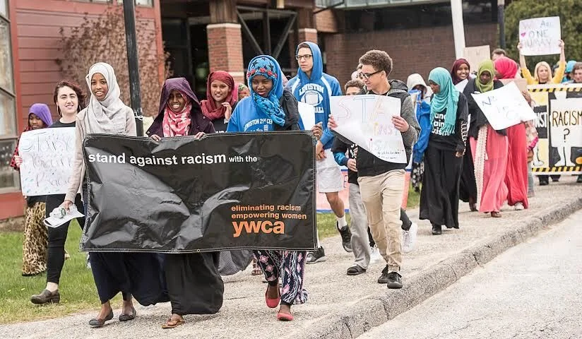 A diverse group of people participating in a march against racism, holding signs and a banner that reads 'stand against racism with the YWCA'.