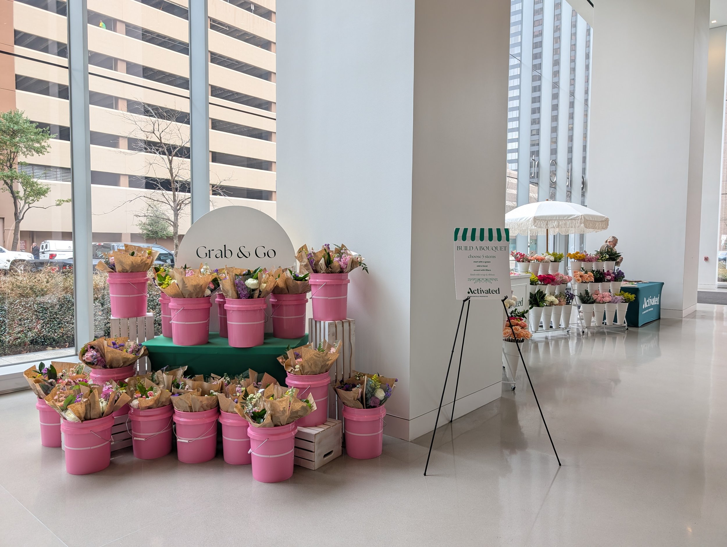 Indoor flower bar display with pink buckets of flowers, a sign that reads 'Grab & Go', and a table with flower wrappers and a person browsing.