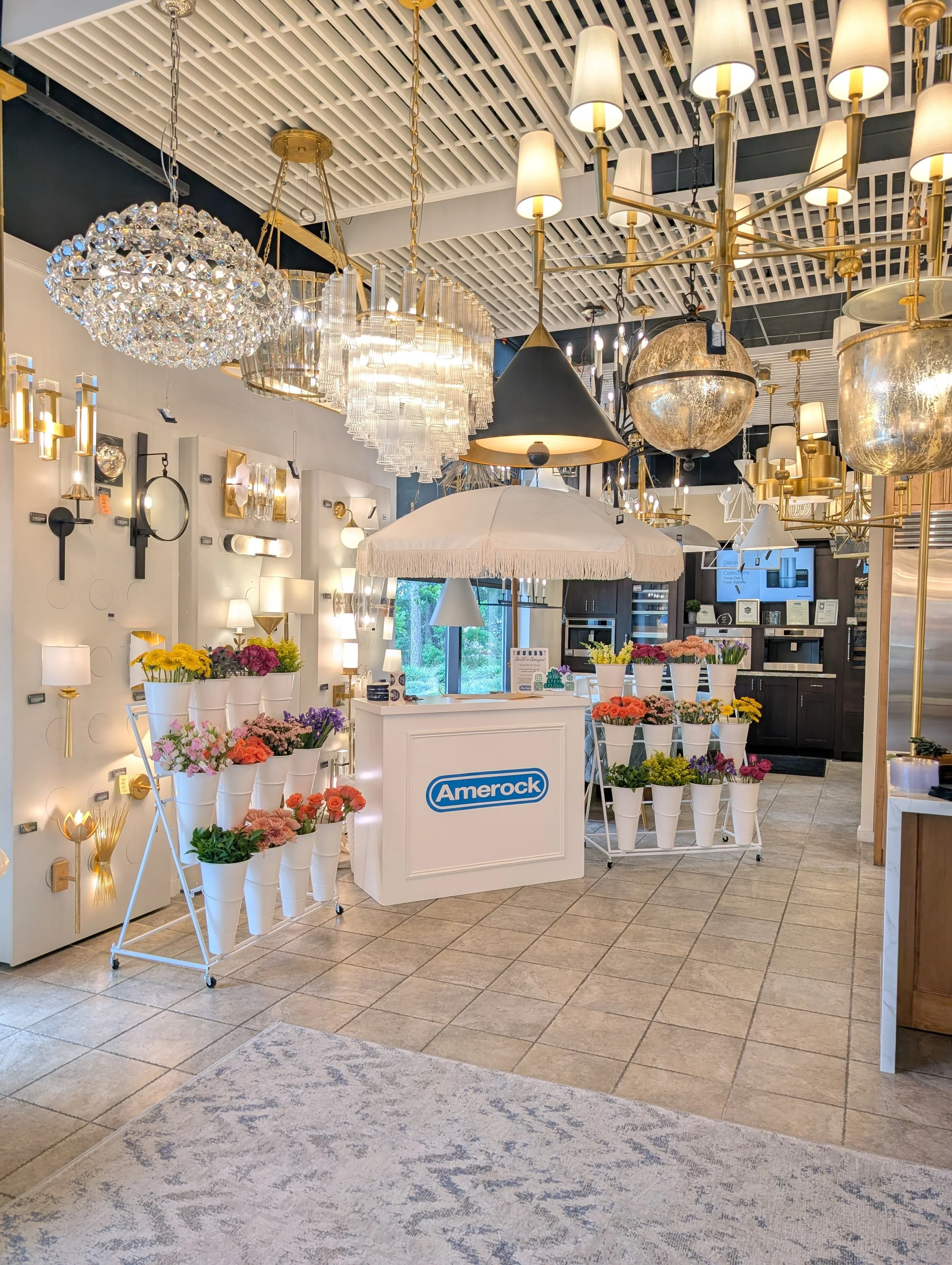 Interior of a lighting store with various chandeliers, wall sconces, and lamps on display, including flowers on a white display stand and a white umbrella.