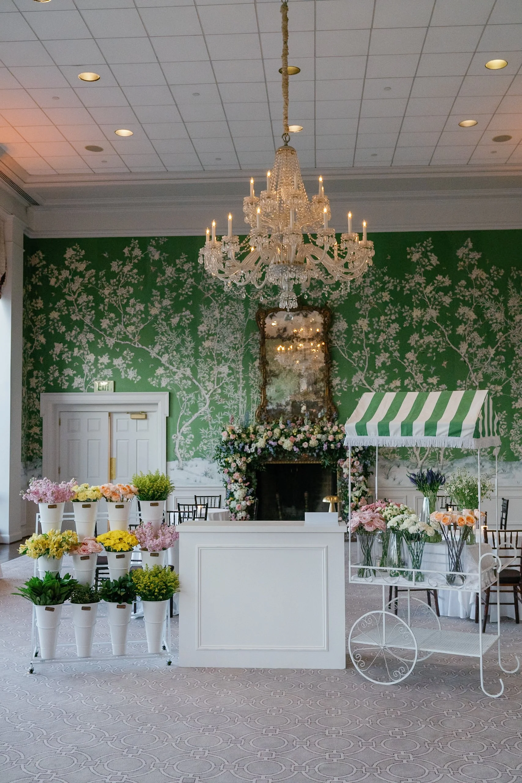 Flower bar setup with bucket stand containing yellow, pink and green florals, a white bar, and a metal cart displaying more flowers set in a grand ballroom with green vintage wallpaper and a large chandelier
