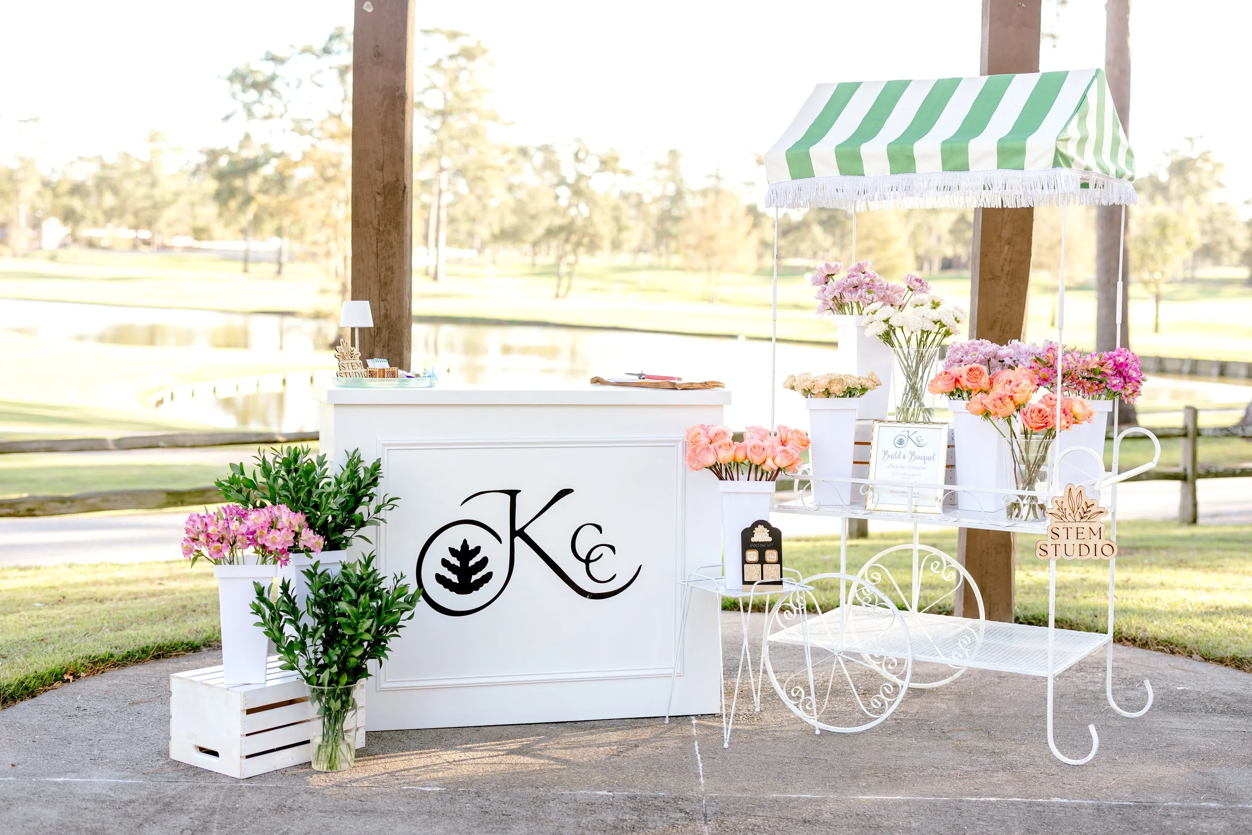 Bouquet bar set up with white bar displaying black KCC logo, and metal cart with green and white stripe top displaying brightly colored florals