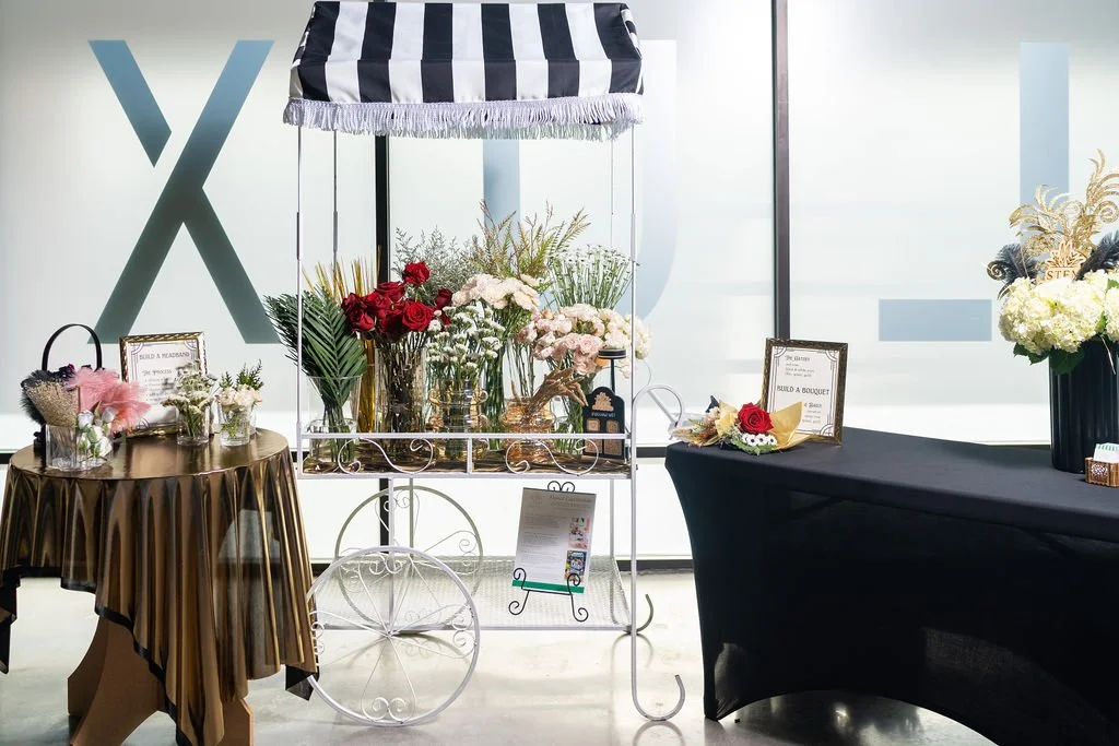 Flower bar setup with small round table covered in gold linen, white metal cart displaying romantic florals