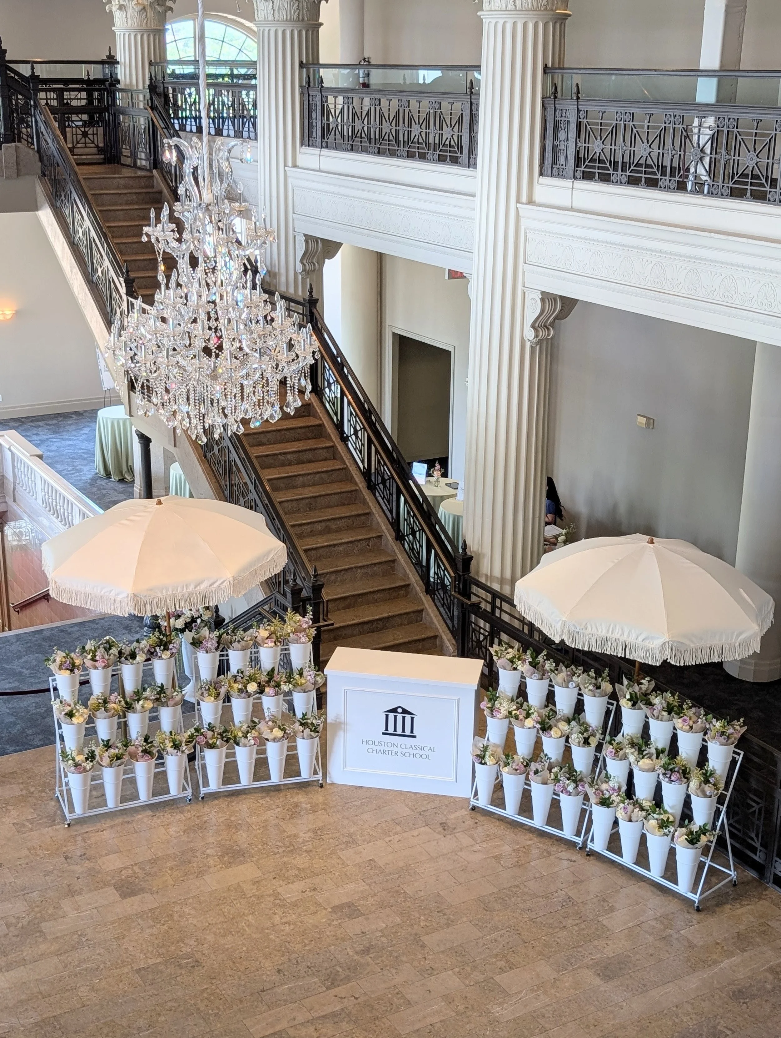 Interior view of a grand hall with a staircase, large chandelier, and decorative columns. There are flower arrangements in bucket stands with white fringe umbrellas and a corporate logo on the decorative bar.
