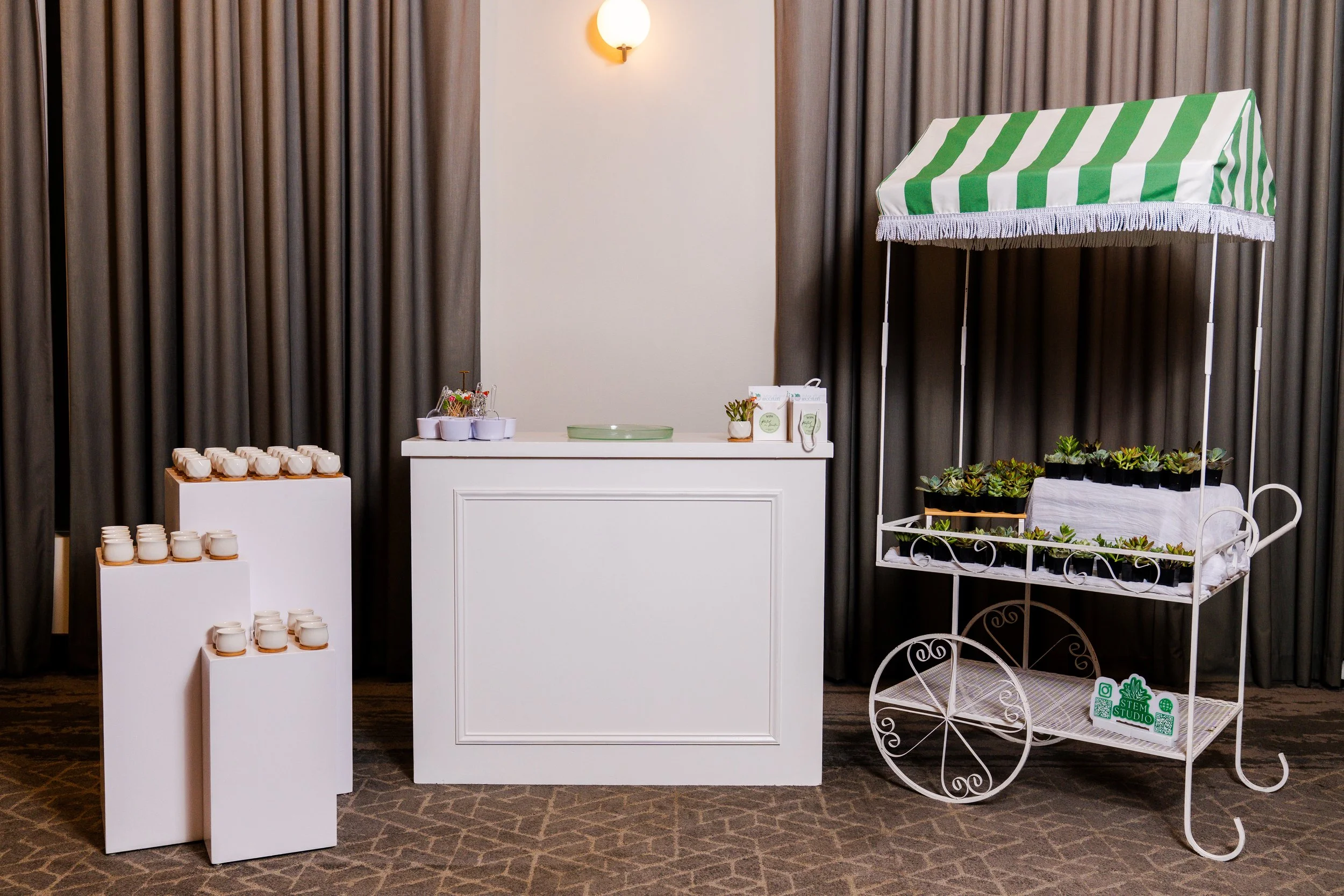 succulent bar setup with three nested white pedestals on left, white bar in center, and flower cart displaying succulent plants with green and white striped top on right