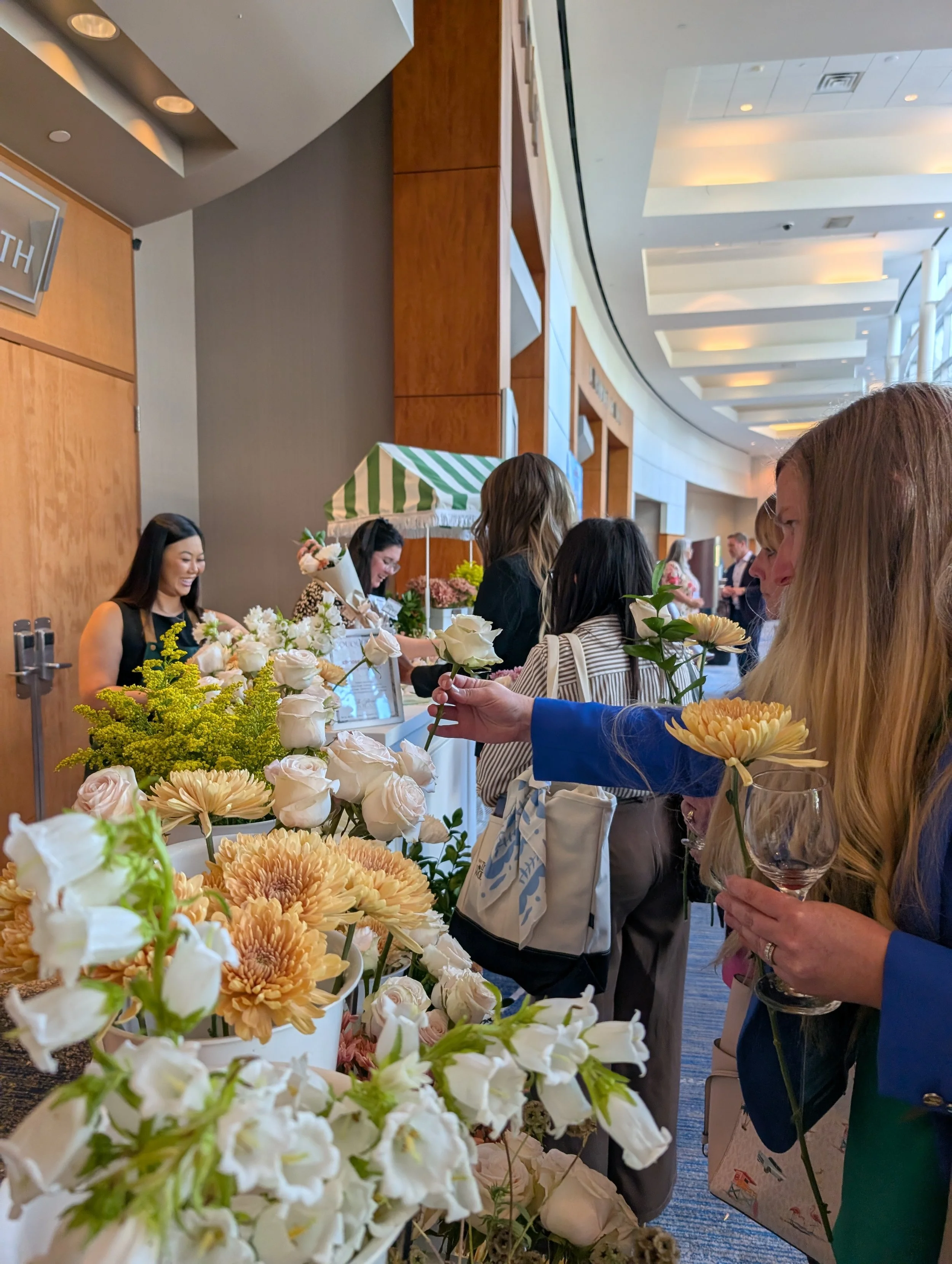 People at a flower stand in a hallway, selecting and designing floral bouquets. The stand displays a variety of flowers including roses, chrysanthemums, and greenery. The background shows a modern interior with wood paneling and ceiling lights.