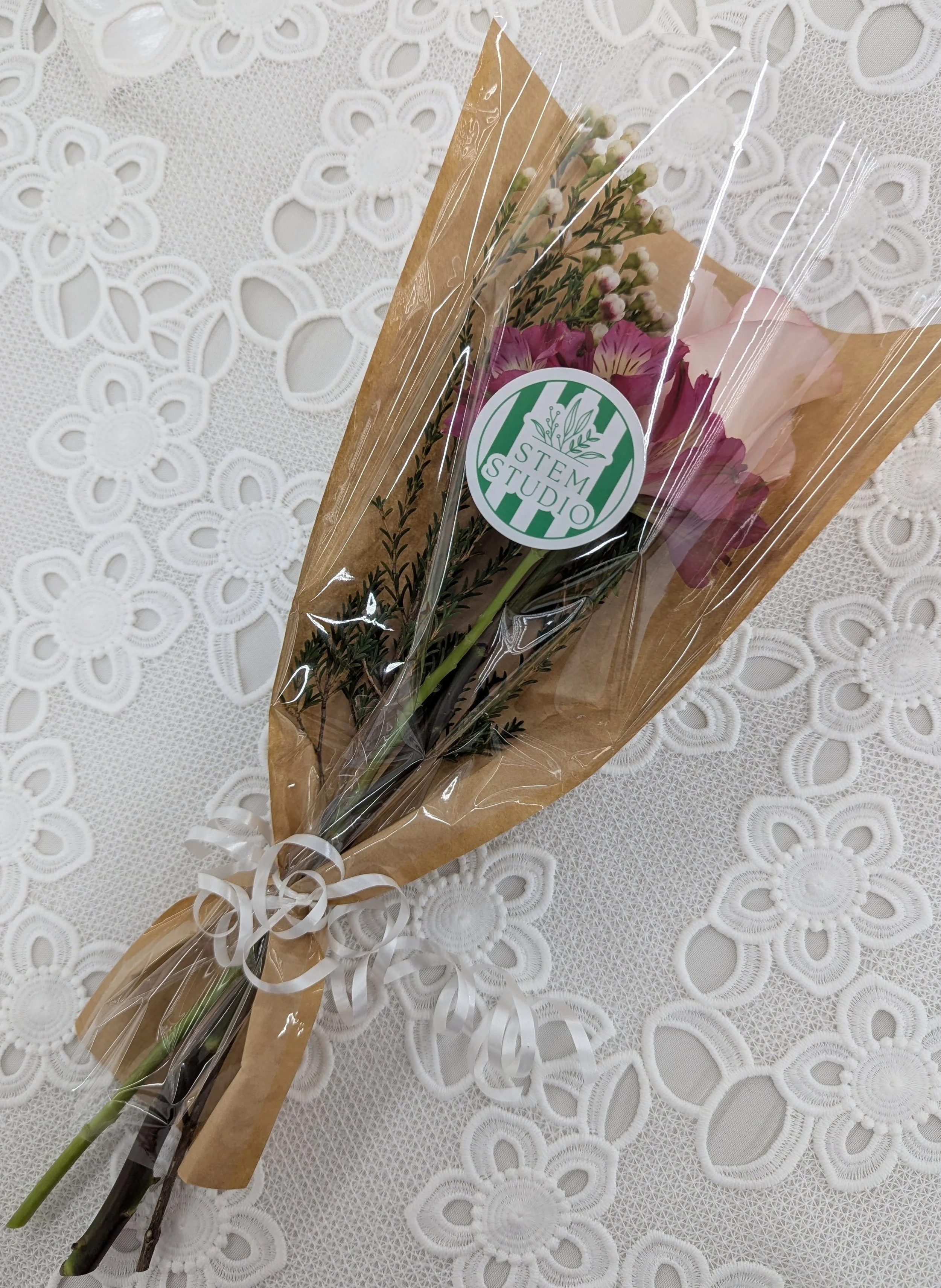 A bouquet of pink and white flowers wrapped in clear plastic with a 'Stem Studio' sticker, placed on a lace tablecloth.