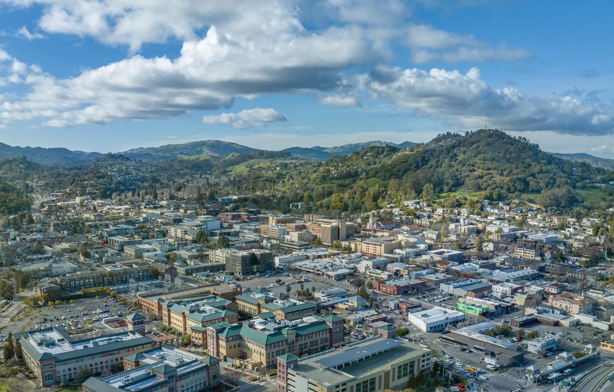 Cityscape with a mix of commercial and residential buildings, surrounding hills covered in trees, partly cloudy sky.