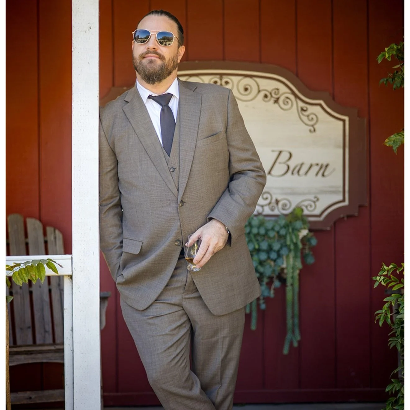 A man in a suit and sunglasses leaning against a white post, holding a glass, in front of a red barn with a decorative sign that reads "Barn".