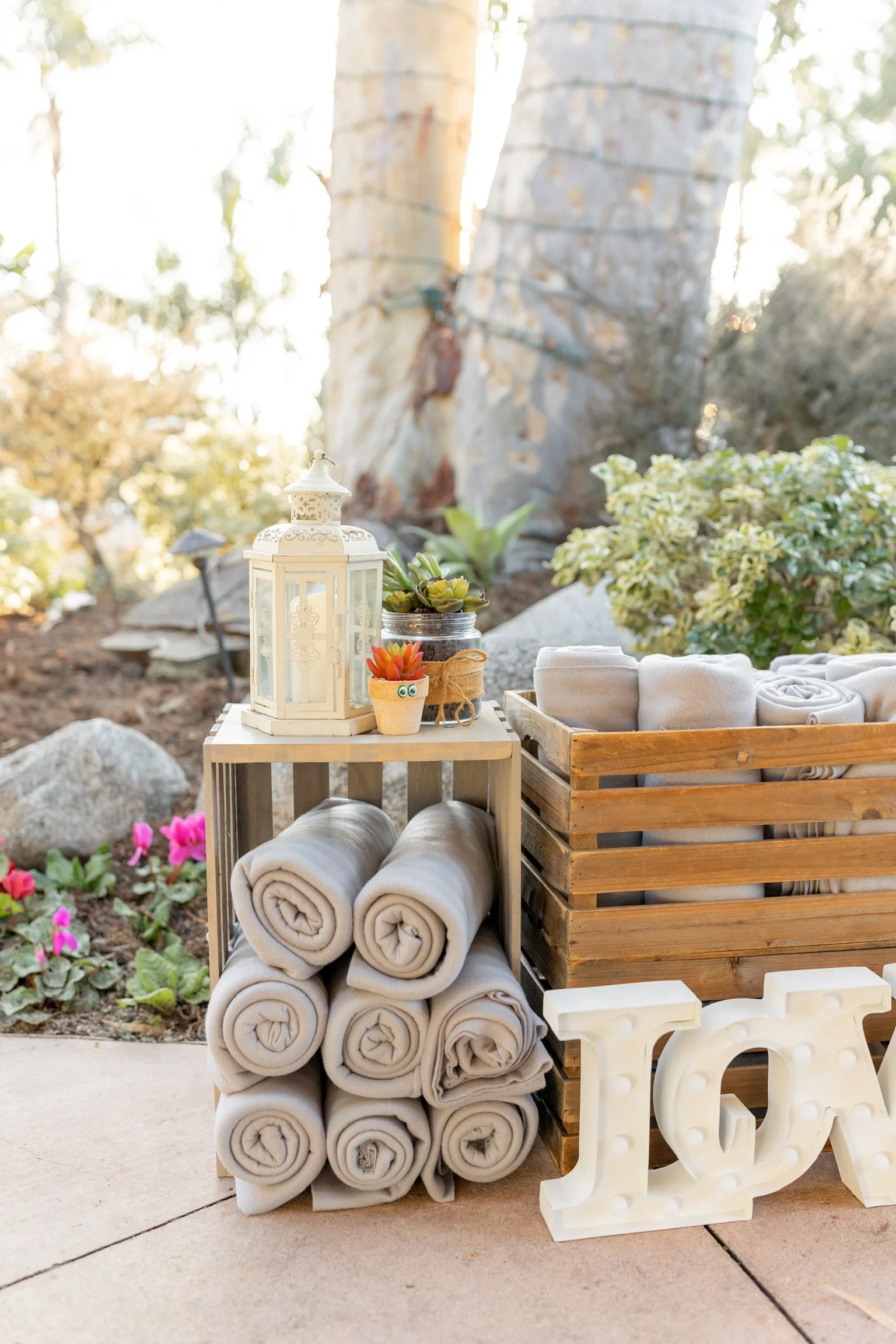 Outdoor scene with rolled beige towels, a white lantern, small potted succulent plants, and a wooden crate, with decorative large white 'LOVE' letters on a paved surface near a garden with colorful flowers and a large tree.