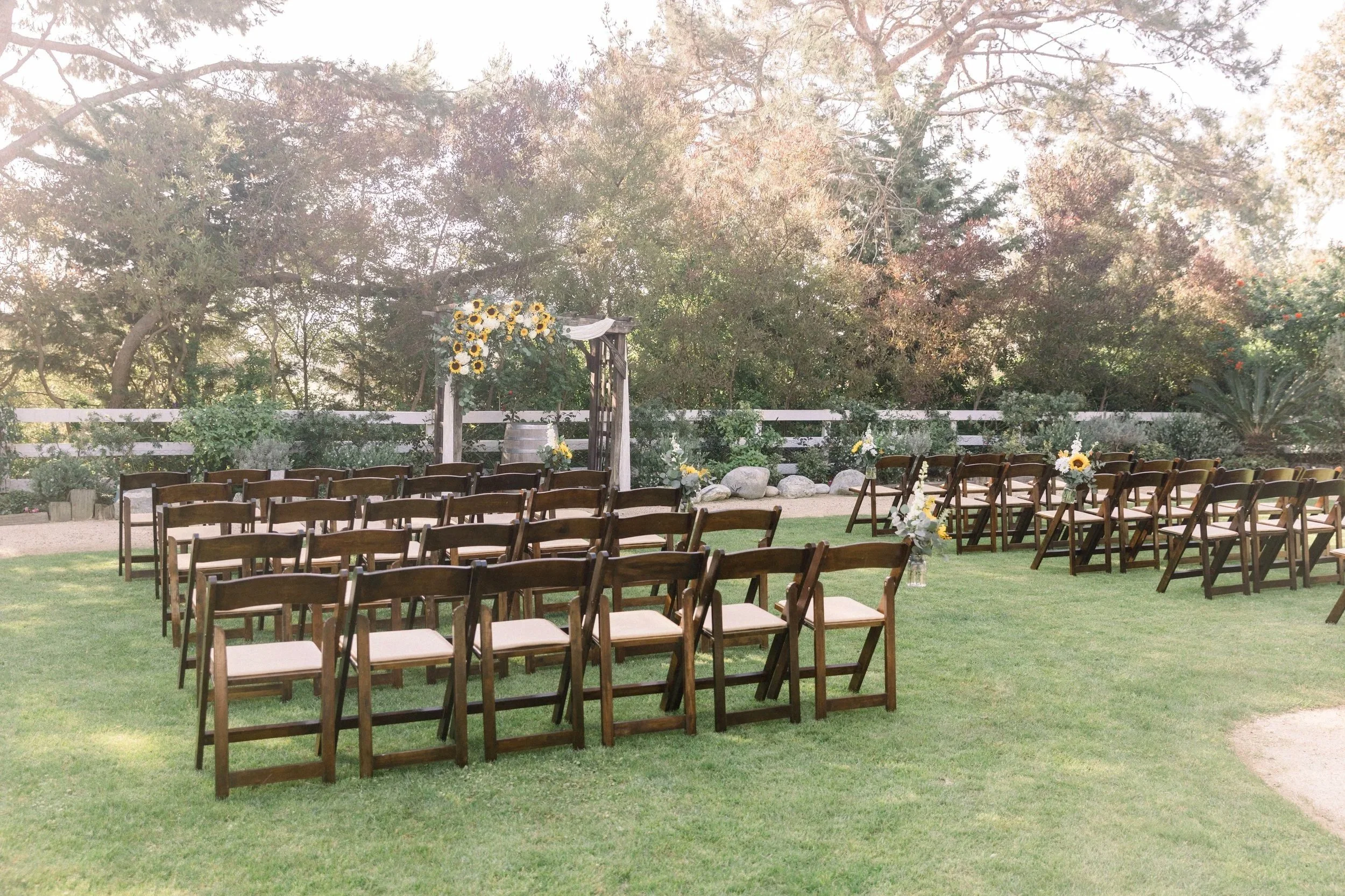 Outdoor wedding ceremony setup with rows of wooden chairs, floral arrangements, and an arch decorated with sunflowers and white drapes, on a green lawn surrounded by trees and shrubs.