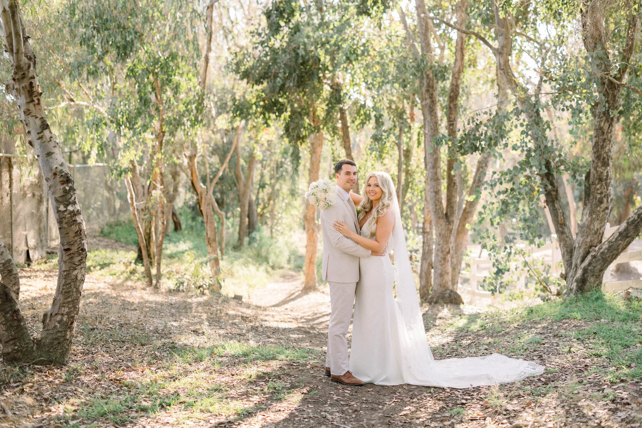 A newlywed couple standing in a wooded outdoor setting with sunlight filtering through the trees, the groom holding a bouquet of white flowers, both dressed in formal wedding attire, embracing and smiling.