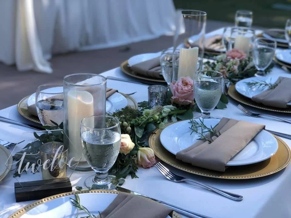 Elegant outdoor dining table set with white tablecloth, gold chargers, white plates, beige napkins with greenery, and glassware, decorated with candles and pink flowers for a formal event.