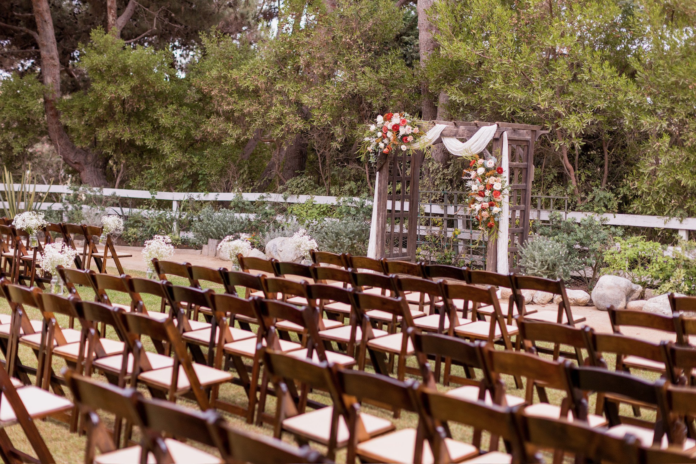 Outdoor wedding ceremony setup with wooden chairs arranged facing a decorated wooden arch, surrounded by trees and greenery.