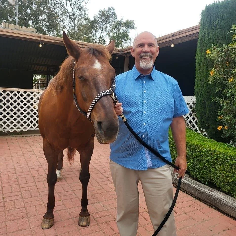 A man with a bald head, gray beard, and mustache holding a horse's reins, standing on a paved area outside a house with white lattice fencing and green bushes.