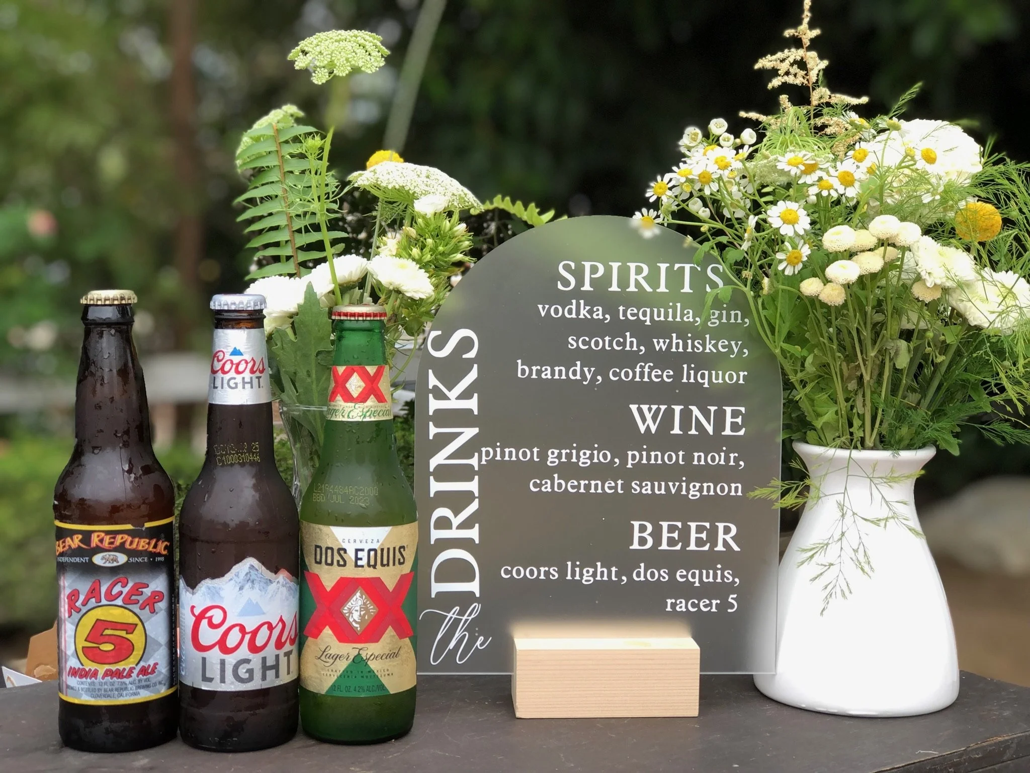 A display of beer bottles and a floral arrangement with a blackboard sign listing different spirits, wines, and beers. The sign mentions vodka, tequila, gin, scotch, whiskey, brandy, coffee liquor, pinot grigio, pinot noir, cabernet sauvignon, Coors Light, Dos Equis, and Racer 5.