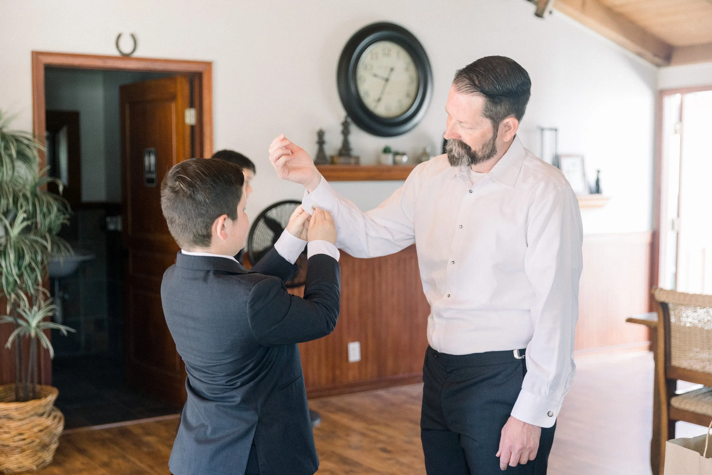 A man adjusts the cuff of a young boy dressed in a suit inside a home decorated with wall clock, plants, and wooden furniture.