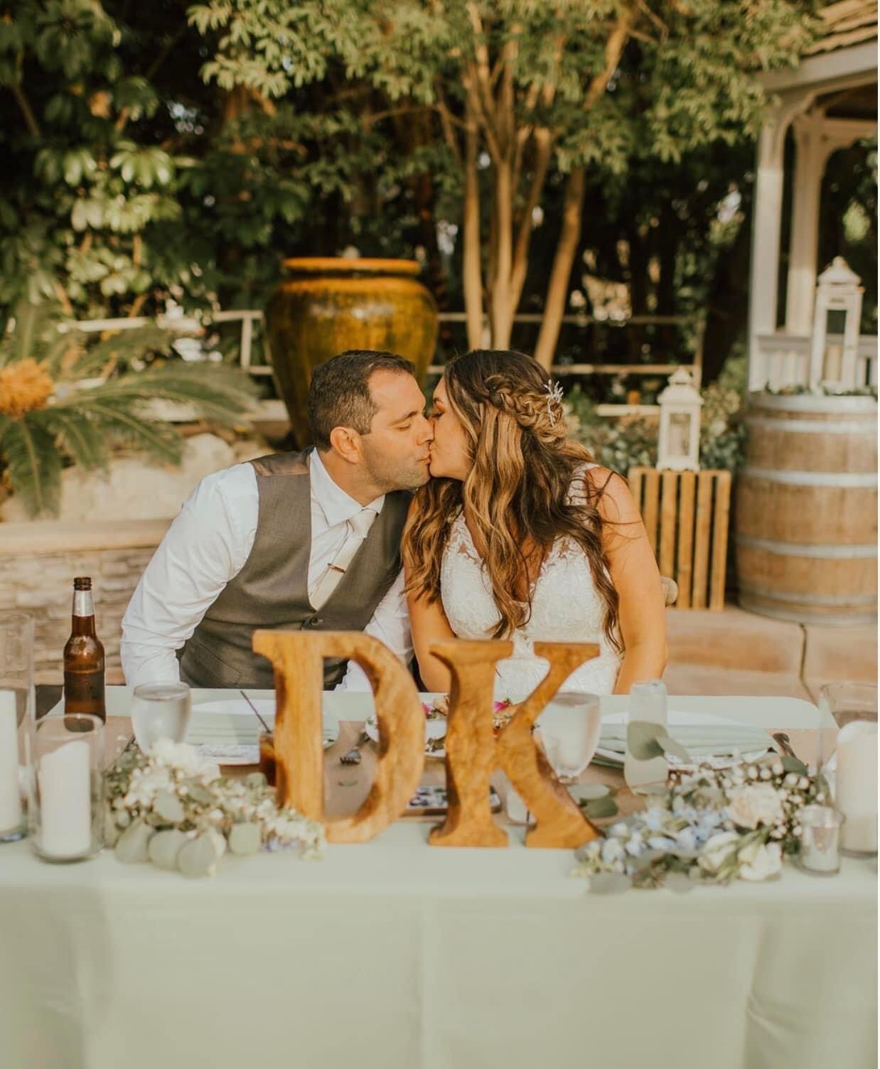 A newlywed couple shares a kiss at their wedding reception table decorated with flowers, candles, and wooden initials D and K.