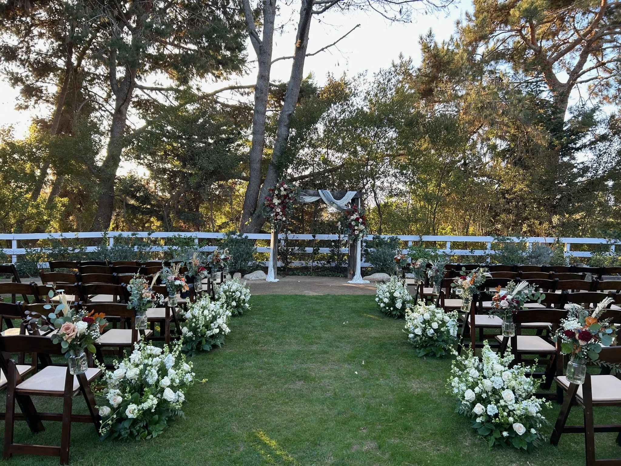 Outdoor wedding setup with chairs arranged along an aisle decorated with white and pink floral arrangements, leading to a wedding arch draped in white fabric and flowers, surrounded by trees and greenery.