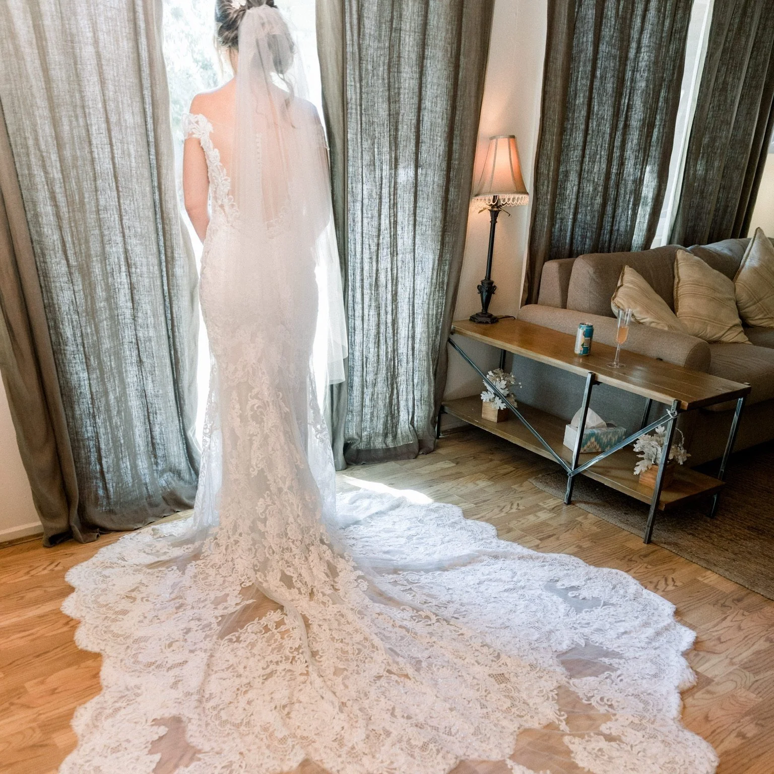A woman in a white wedding dress with lace details, standing behind semi-sheer curtains in a living room, with sunlight shining through.