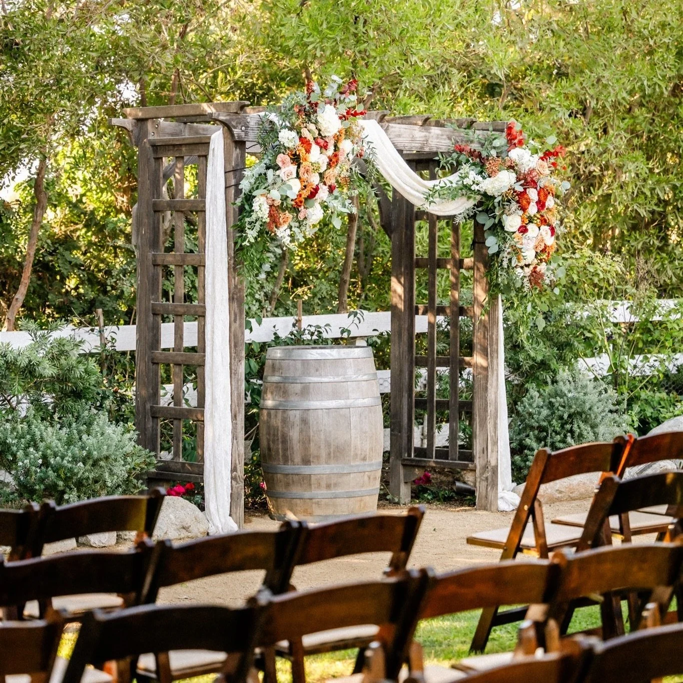Outdoor wedding ceremony setup with a wooden arch decorated with white, pink, and red flowers and greenery, a wooden barrel beneath the arch, and folding chairs for guests in a garden setting.