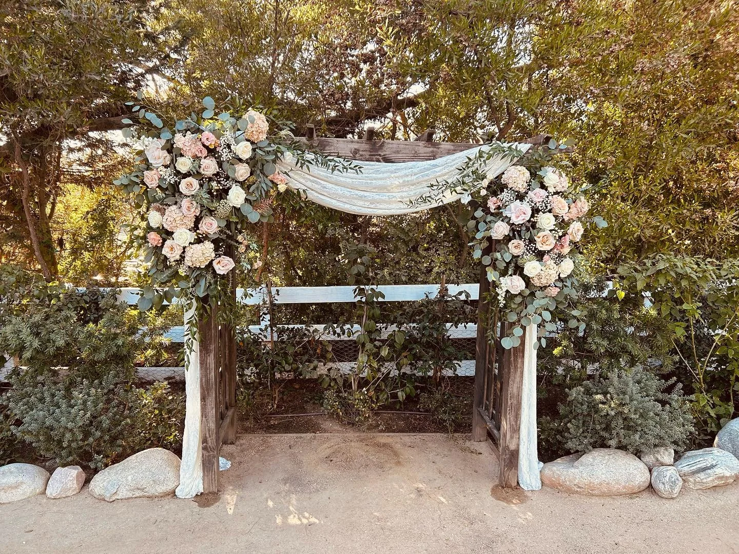 Wedding arch decorated with white and blush pink flowers and greenery, draped with white fabric, in an outdoor garden setting.