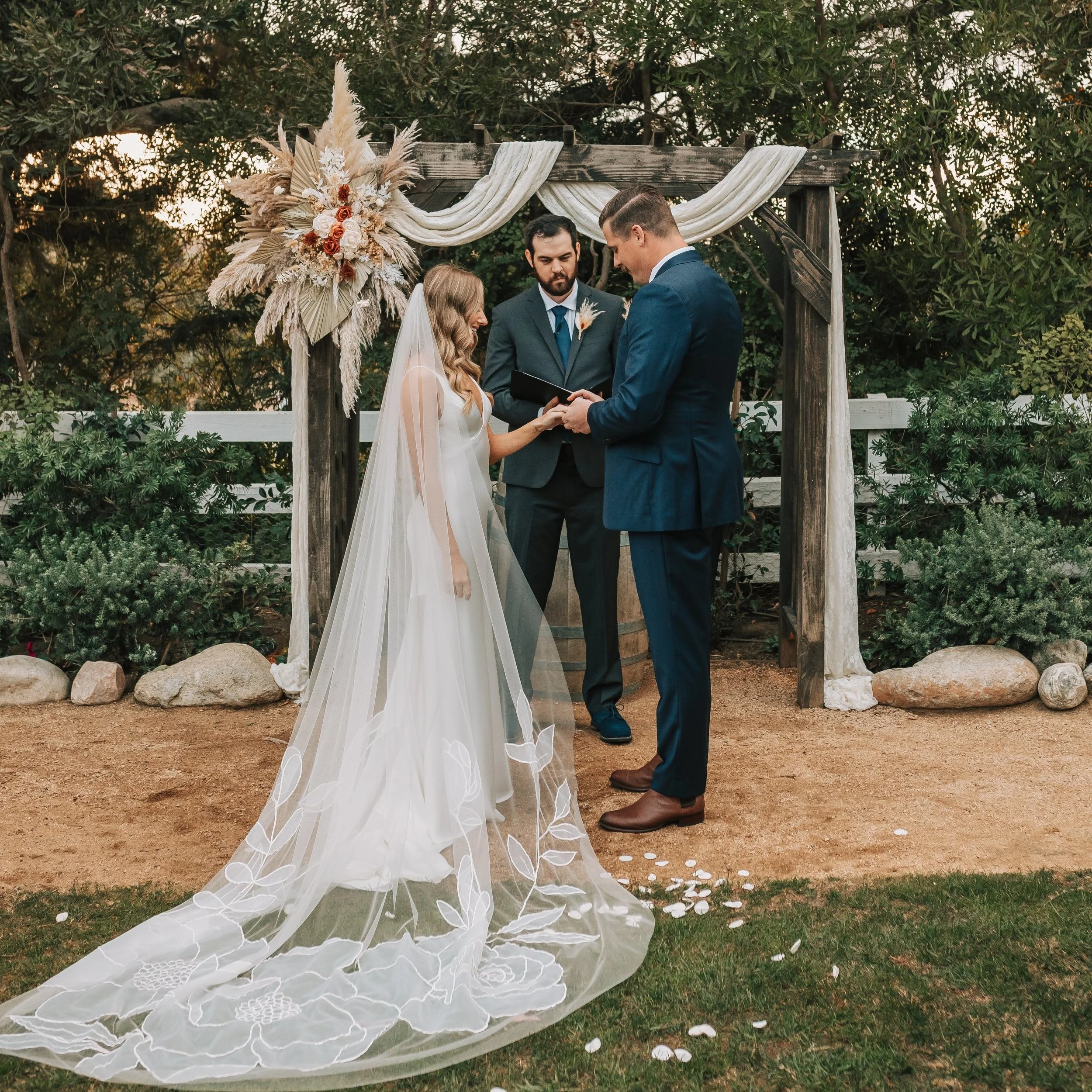 A couple getting married outdoors, standing under a wooden wedding arch decorated with dried flowers and fabric drapes. The bride wears a white dress with a long, embroidered veil, and the groom wears a dark blue suit. An officiant stands behind them, reading from a book. Flower petals are scattered on the ground in front of them.