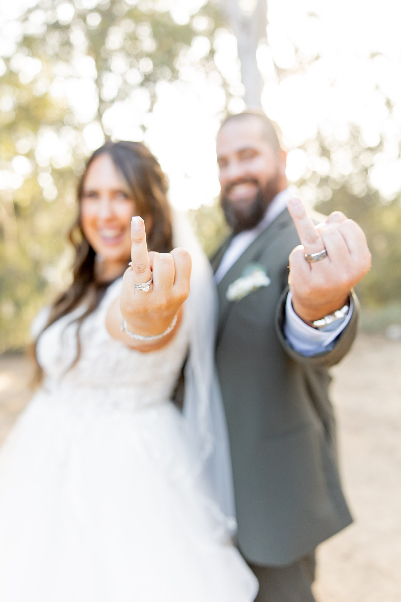 A newlywed couple outdoors showing their wedding rings while making a rude gesture with their middle fingers.