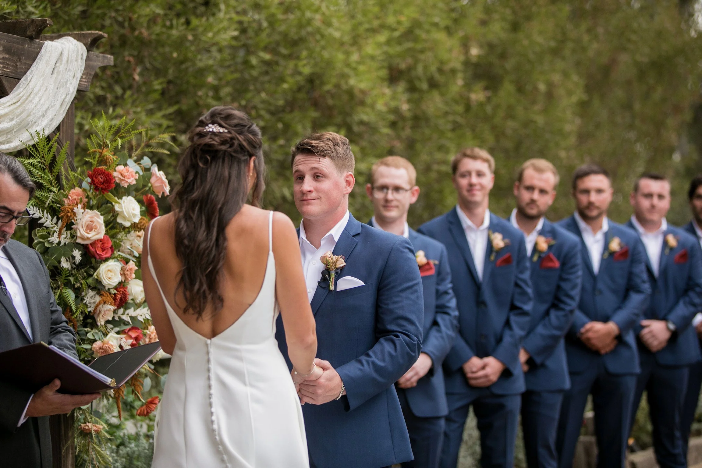 A bride and groom at Red Horse Barn exchange vows during an outdoor wedding ceremony, with groomsmen in blue suits standing behind them and floral decorations on the side.