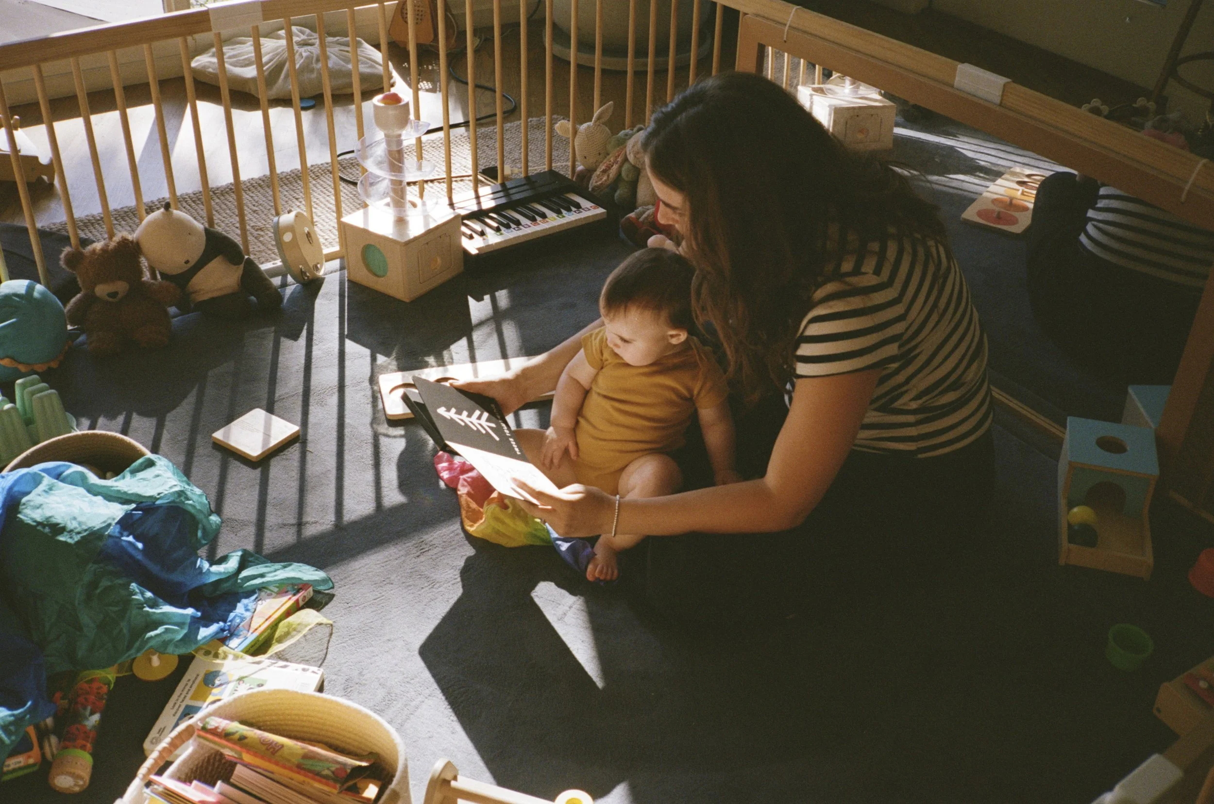 A woman and a young boy sitting on the floor of a toddler's play area, reading a book together. The play area contains toys, stuffed animals, a small keyboard, and various other items scattered around.